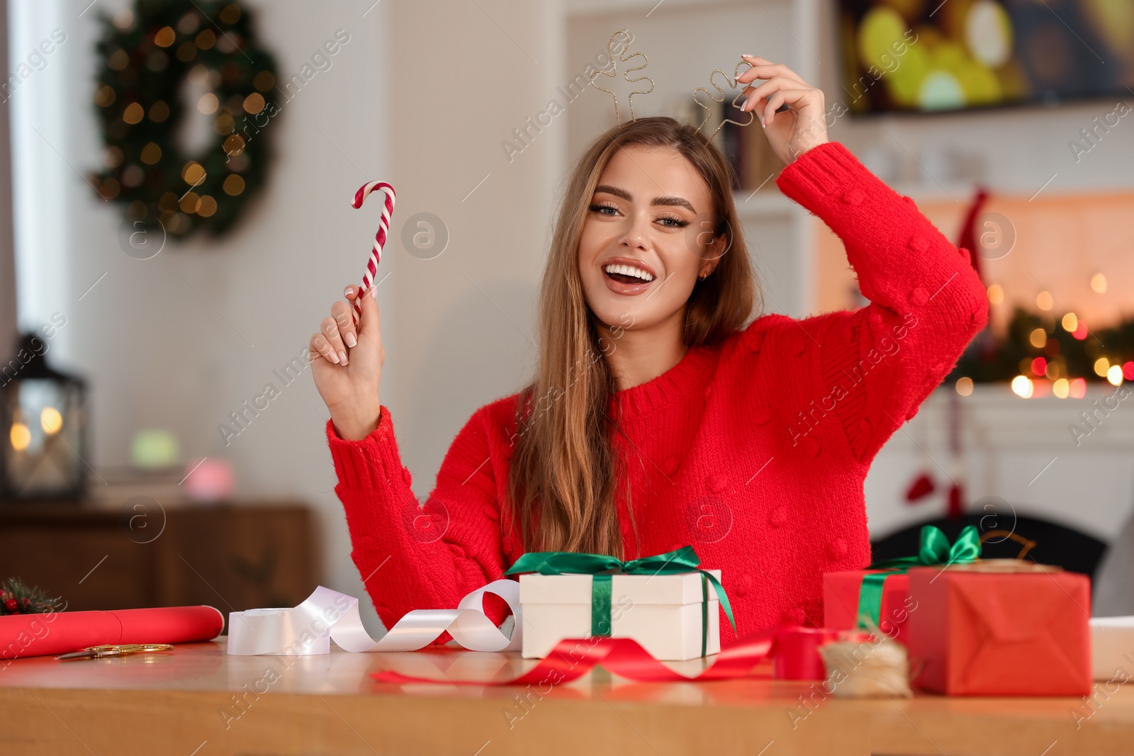 Photo of Beautiful young woman in deer headband with candy cane and Christmas gifts at table in room