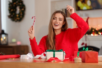 Beautiful young woman in deer headband with candy cane and Christmas gifts at table in room