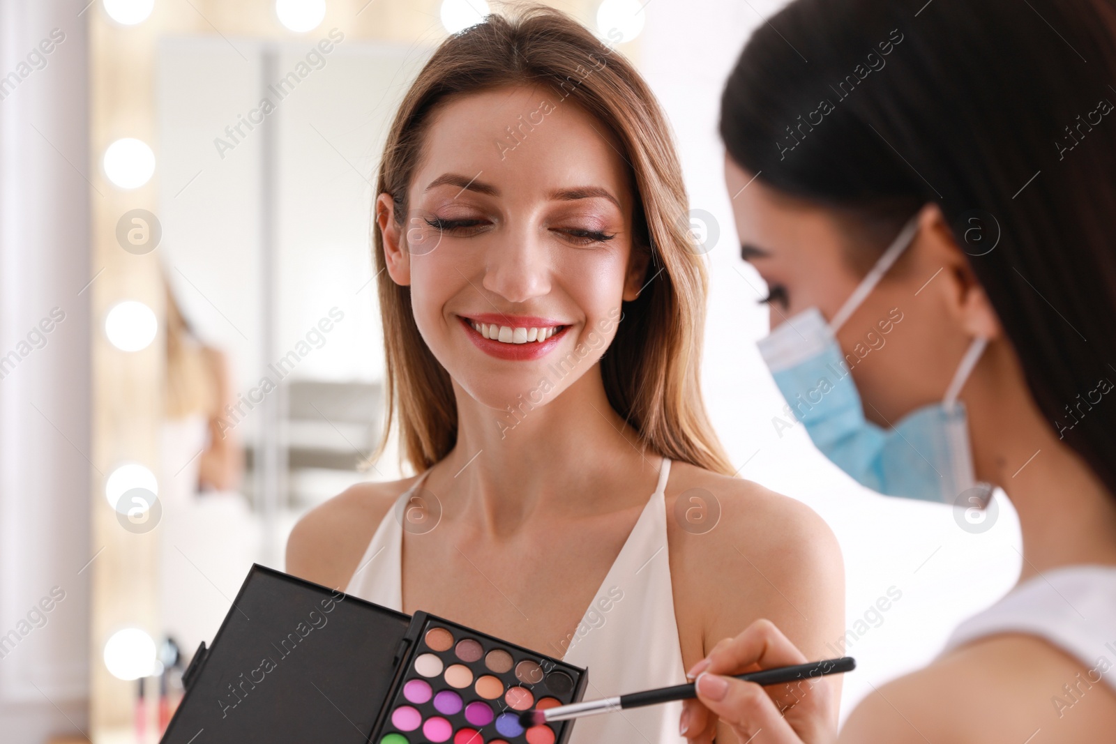 Photo of Makeup artist in protective mask working with woman indoors. Preventive measures during COVID-19 pandemic