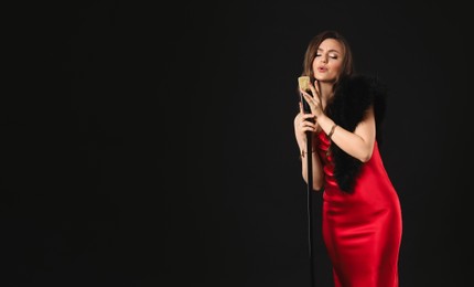 Beautiful young woman in stylish dress with microphone singing on black background