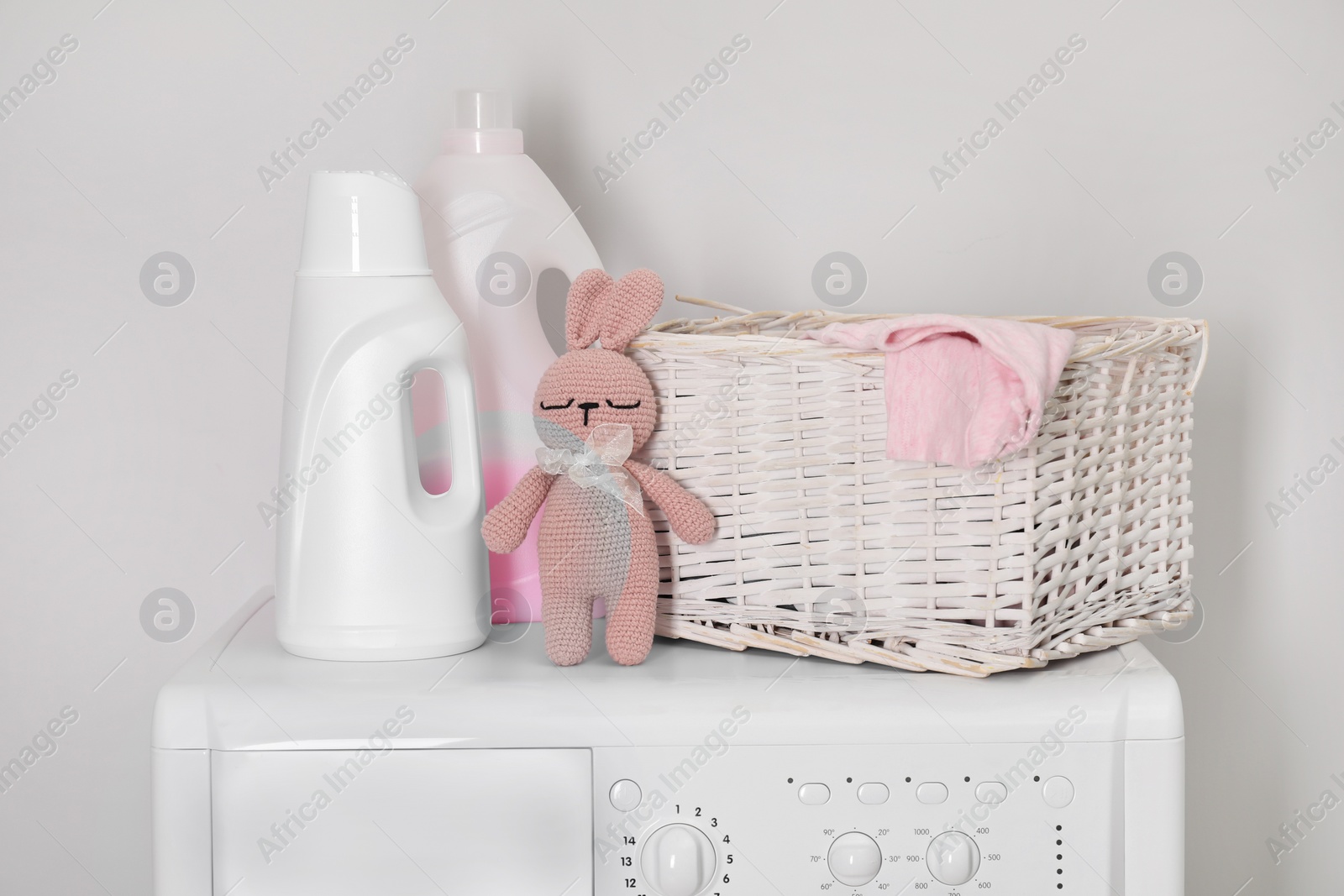 Photo of Baby clothes in wicker basket, laundry detergents and toy bunny on washing machine near light wall