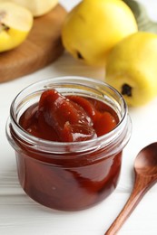 Tasty homemade quince jam in jar, spoon and fruits on white wooden table, closeup