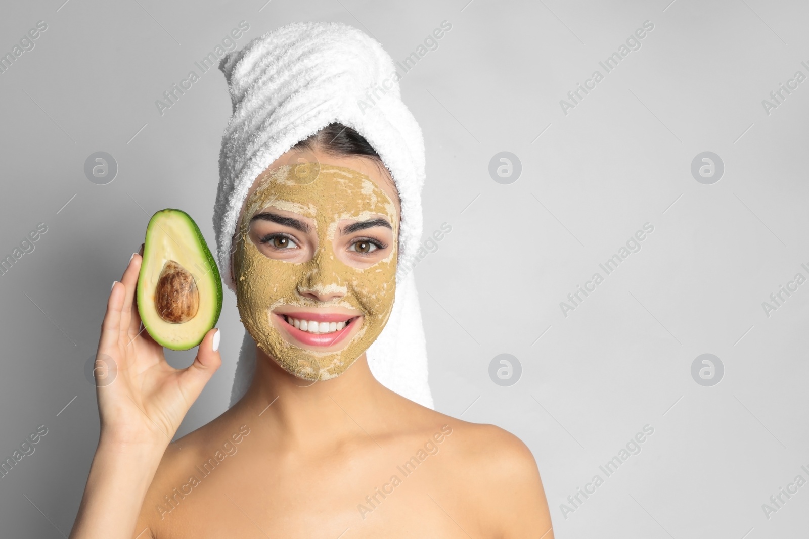 Photo of Young woman with clay mask on her face holding avocado against light background, space for text. Skin care