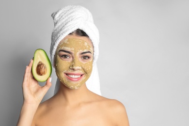 Photo of Young woman with clay mask on her face holding avocado against light background, space for text. Skin care