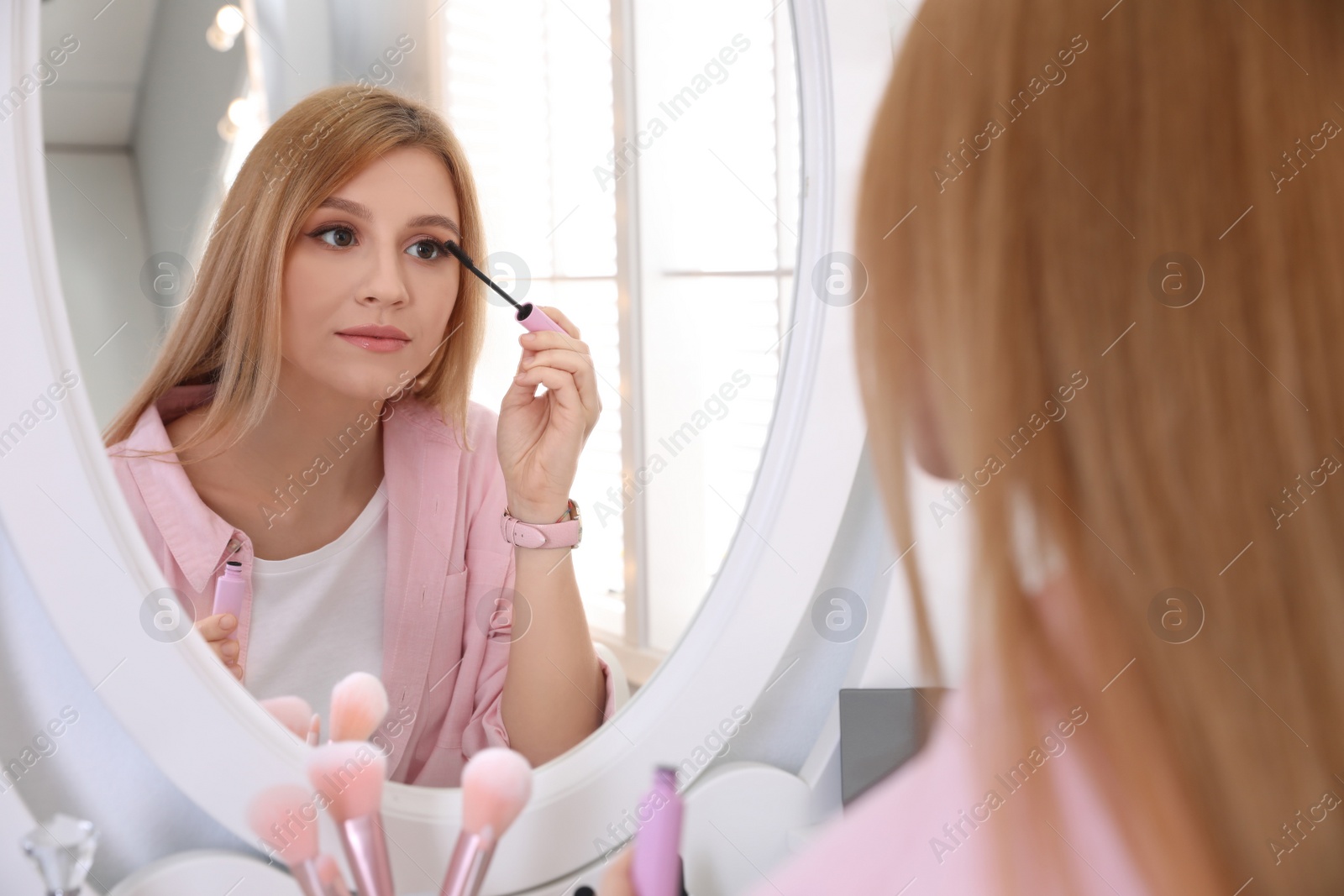 Photo of Beautiful young woman applying makeup near mirror indoors