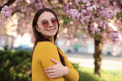 Beautiful woman in sunglasses near blossoming tree on spring day, space for text