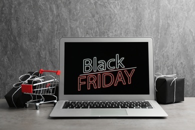 Photo of Laptop with Black Friday announcement, small shopping cart and gifts on table against grey background