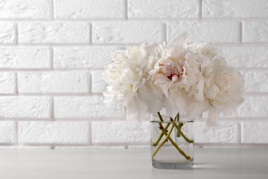Photo of Beautiful peonies in glass vase on white table near brick wall. Space for text