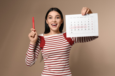 Young woman holding calendar with marked menstrual cycle days on beige background