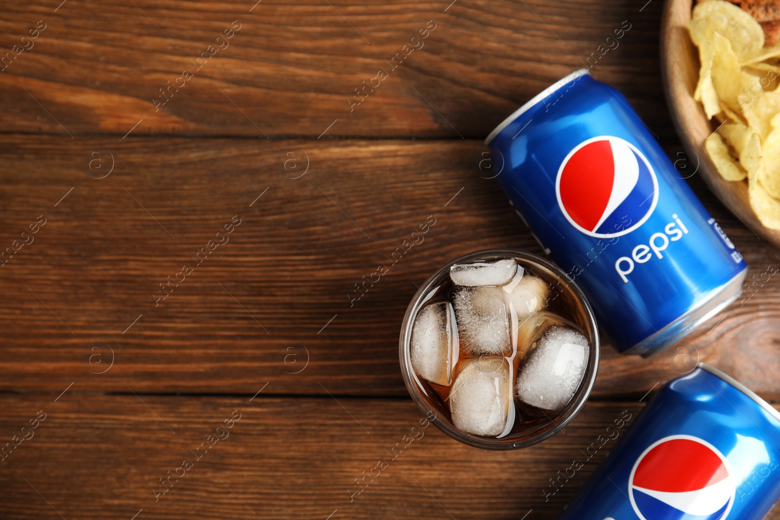 Photo of MYKOLAIV, UKRAINE - FEBRUARY 15, 2021: Glass of Pepsi, cans and snack on wooden table, flat lay. Space for text