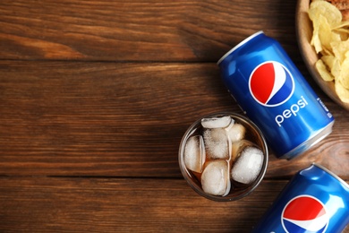 MYKOLAIV, UKRAINE - FEBRUARY 15, 2021: Glass of Pepsi, cans and snack on wooden table, flat lay. Space for text