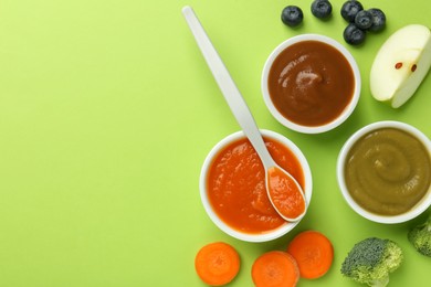 Bowls with healthy baby food, vegetables, fruits and spoon on light green background, flat lay. Space for text