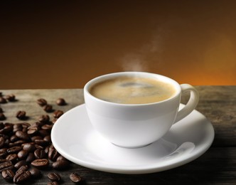 Cup of hot aromatic coffee and roasted beans on wooden table against brown background