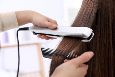 Hairdresser using modern flat iron to style client's hair in salon, closeup