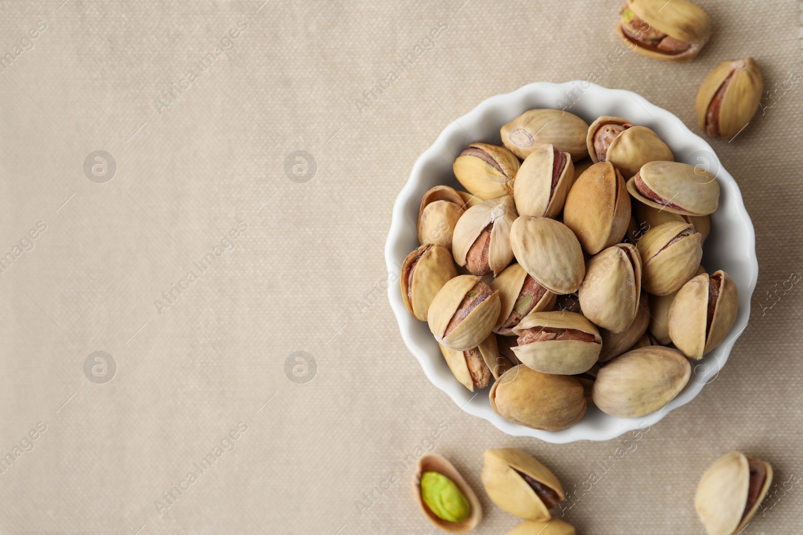 Photo of Bowl and pistachio nuts on beige tablecloth, flat lay. Space for text