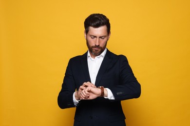 Handsome bearded man in suit looking at wristwatch on orange background