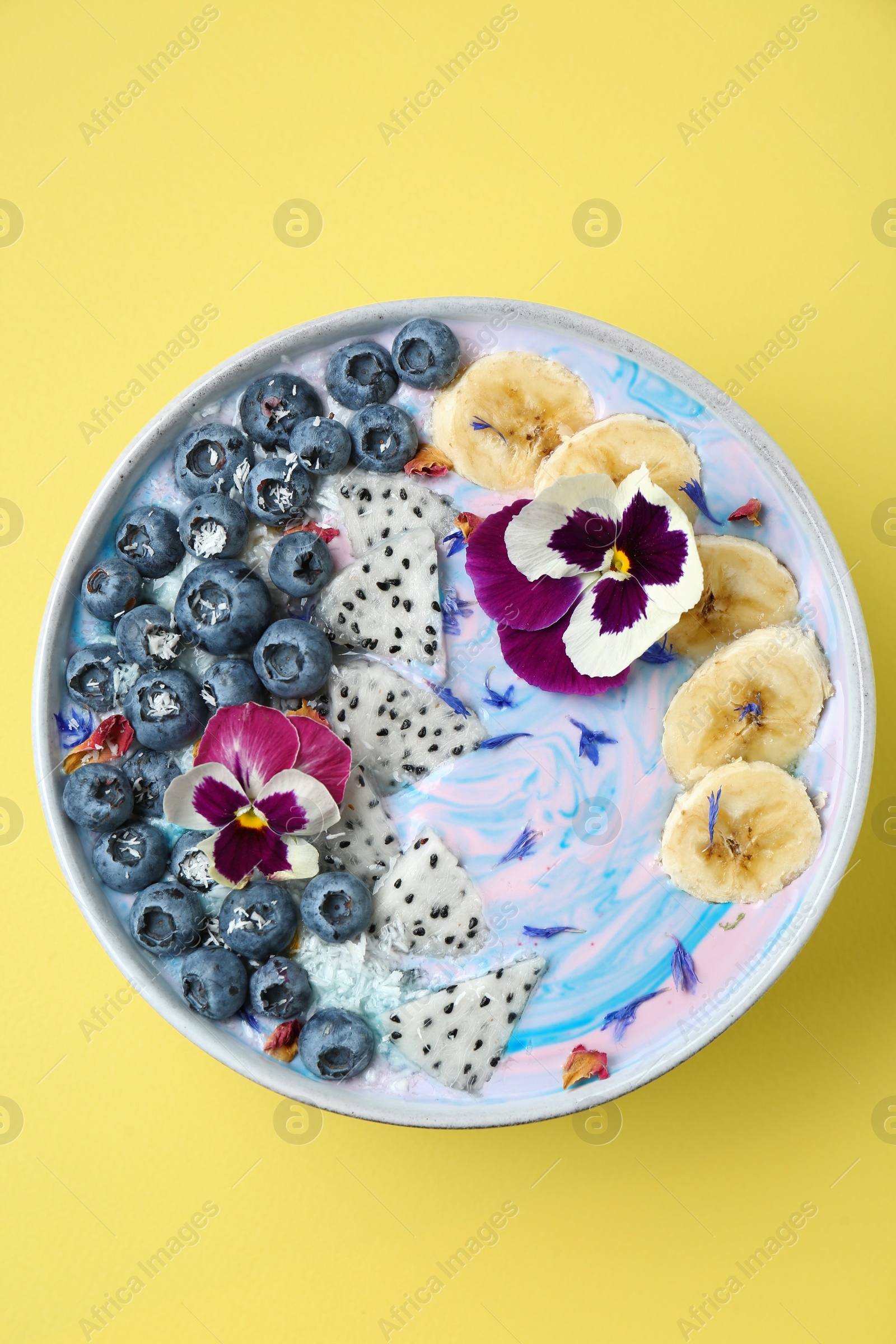 Photo of Delicious smoothie bowl with fresh fruits, blueberries and flowers on yellow background, top view