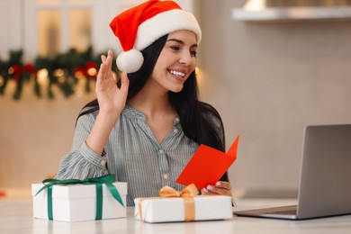Photo of Celebrating Christmas online with exchanged by mail presents. Smiling woman in Santa hat with gifts waving hello during video call at home