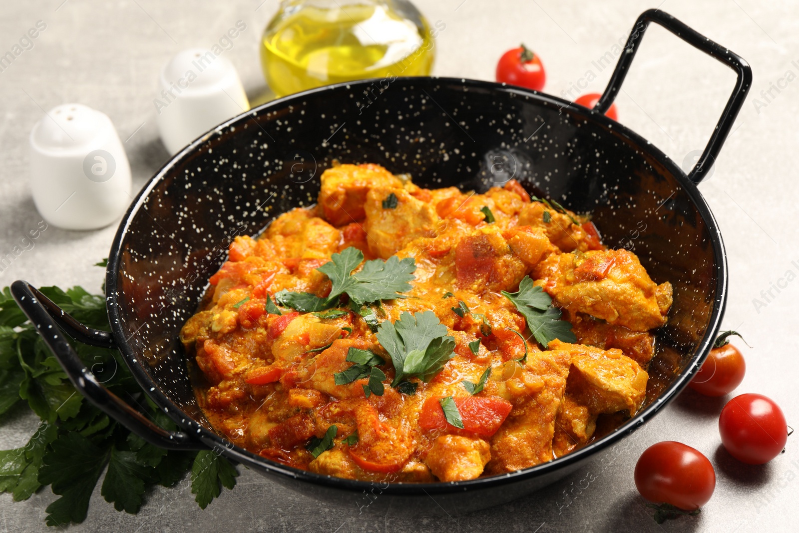 Photo of Delicious chicken curry in frying pan, parsley and tomatoes on light grey table, closeup