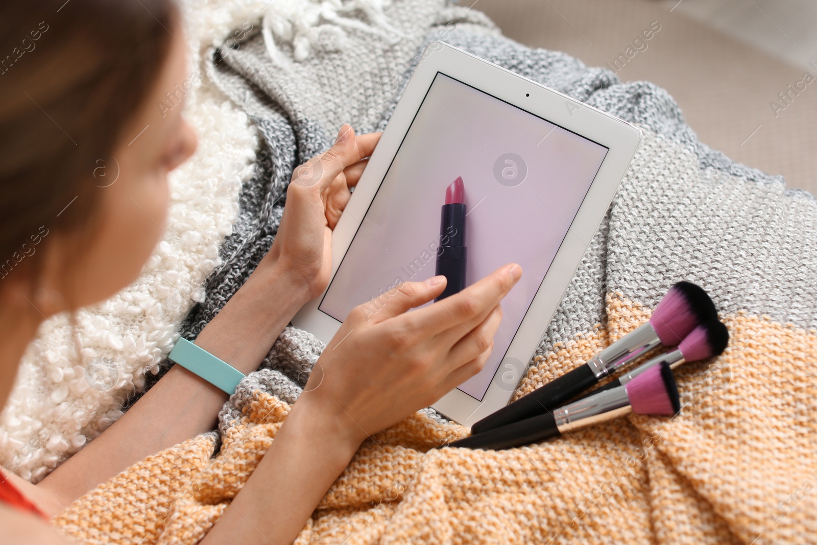 Photo of Young woman with makeup brushes using tablet on bed. Beauty blogger