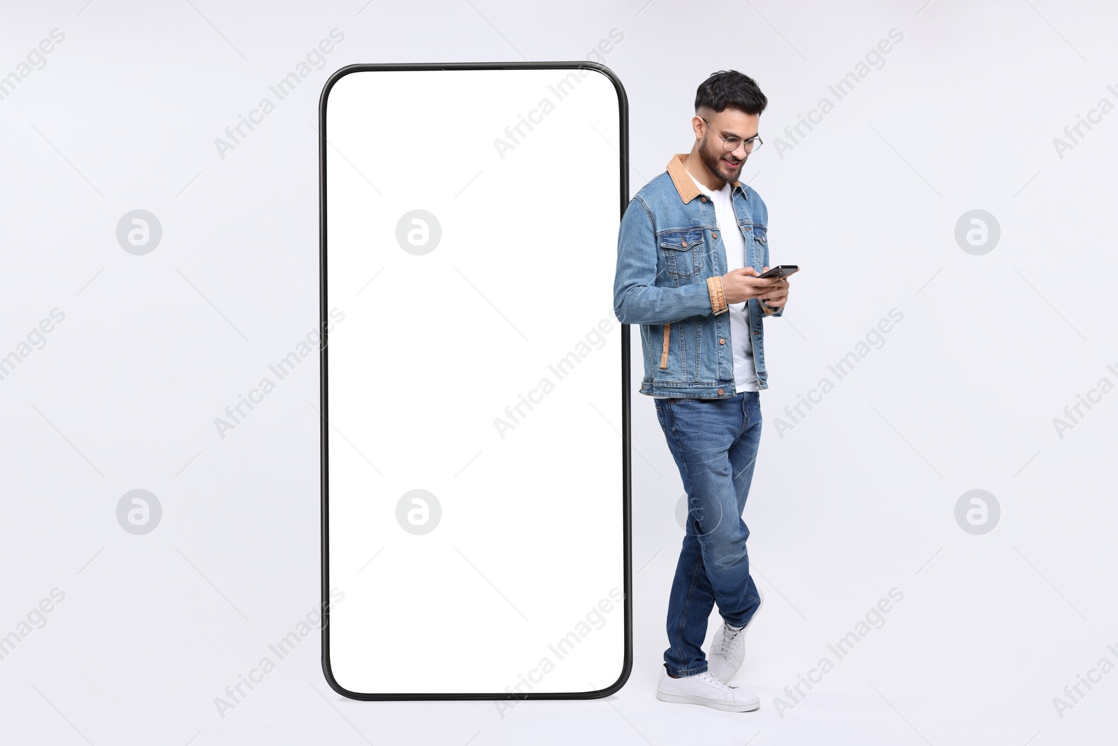 Image of Man with mobile phone standing near huge device with empty screen on grey background. Mockup for design