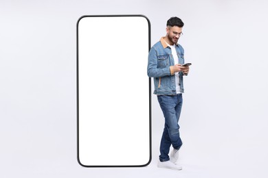 Image of Man with mobile phone standing near huge device with empty screen on grey background. Mockup for design