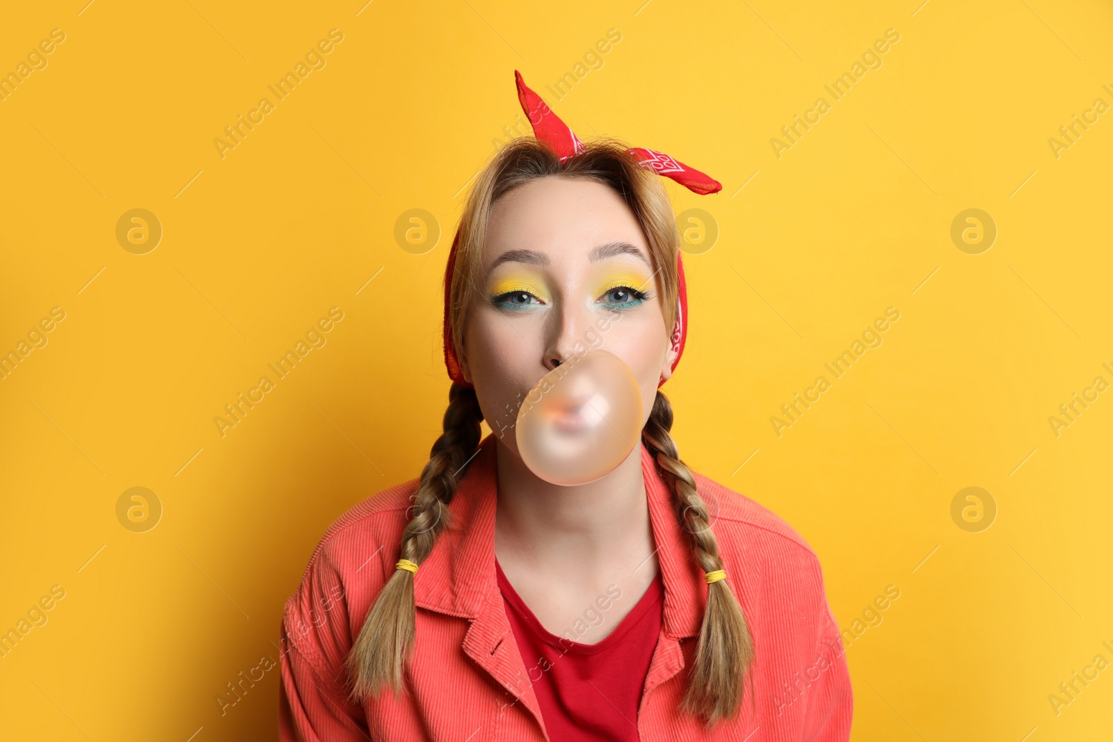 Photo of Fashionable young woman with braids and bright makeup blowing bubblegum on yellow background