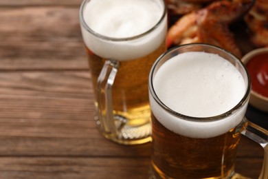Photo of Mugs with beer and delicious baked chicken wings on wooden table, closeup. Space for text