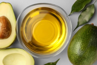 Cooking oil in bowl and fresh avocados on light grey background, flat lay