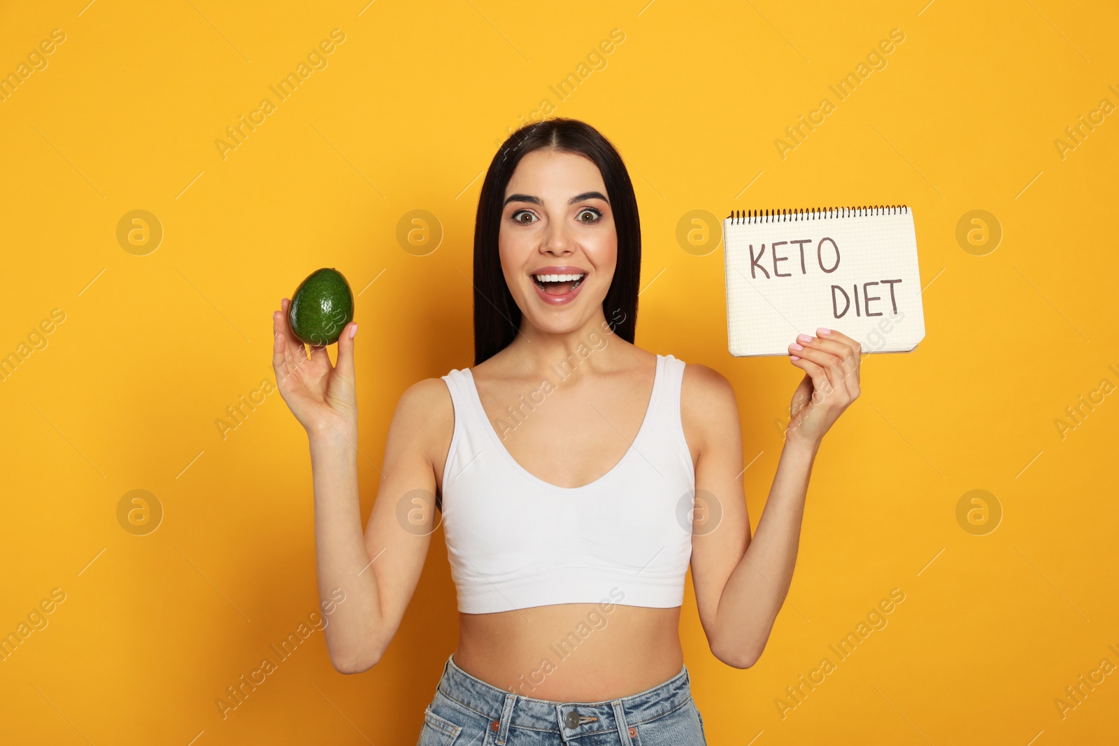 Photo of Happy woman holding avocado and notebook with words Keto Diet on yellow background