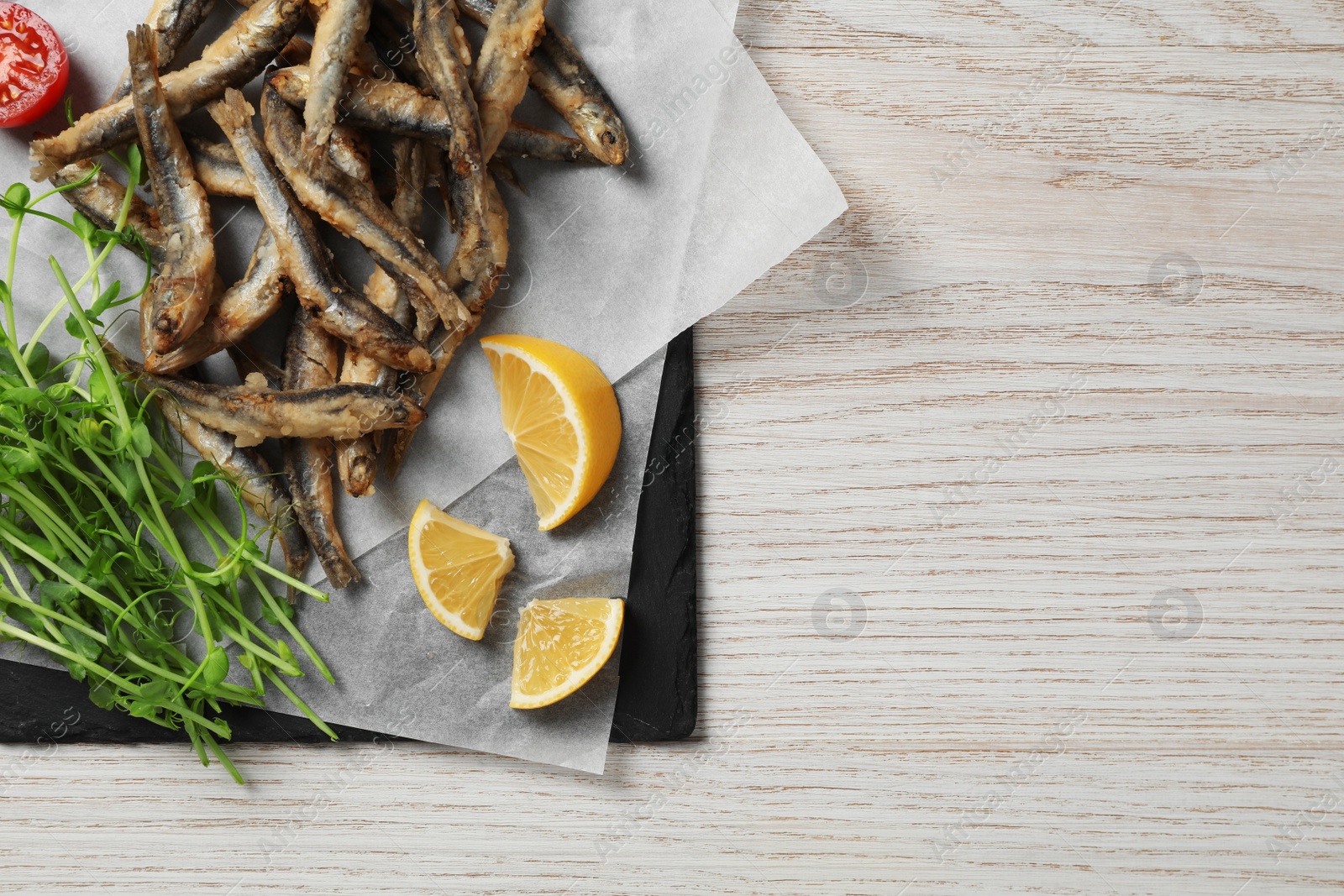 Photo of Slate board with delicious fried anchovies, lime and tomatoes on white wooden table, top view. Space for text