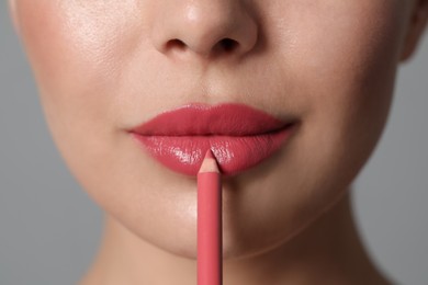 Young woman applying beautiful pink lip pencil on grey background, closeup