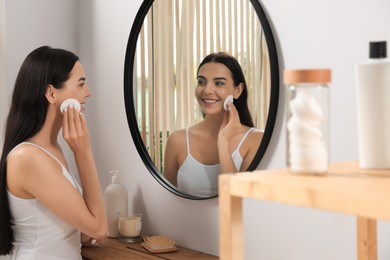 Photo of Young woman using cotton pad with micellar water near mirror indoors