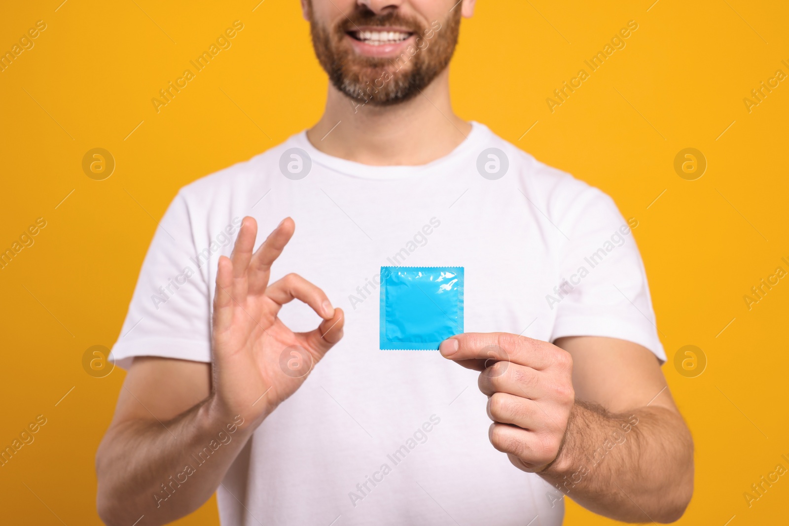 Photo of Man with condom showing ok gesture on yellow background, closeup. Safe sex