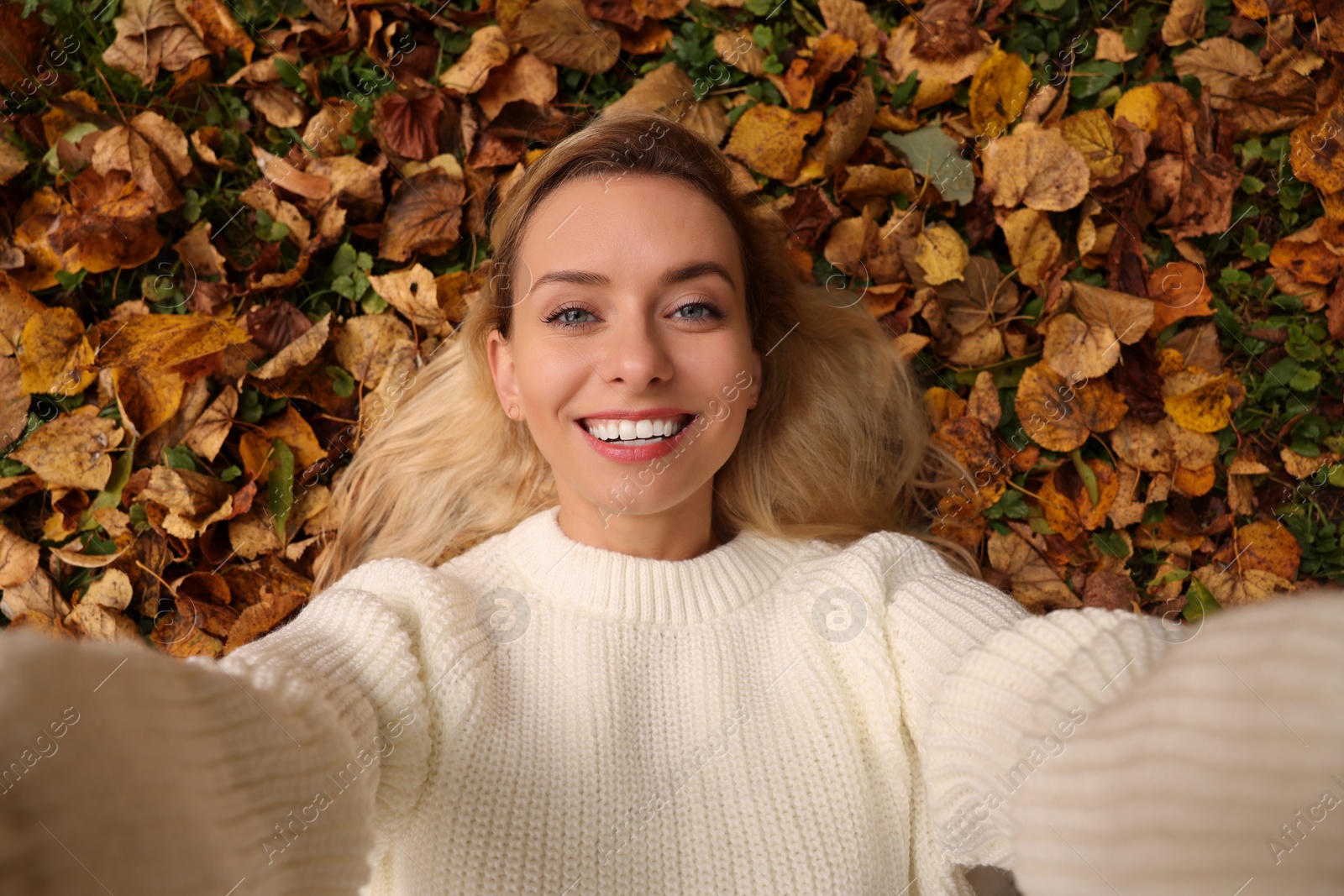 Photo of Smiling woman lying among autumn leaves and taking selfie outdoors, top view