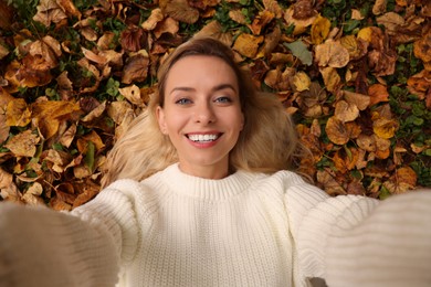 Smiling woman lying among autumn leaves and taking selfie outdoors, top view