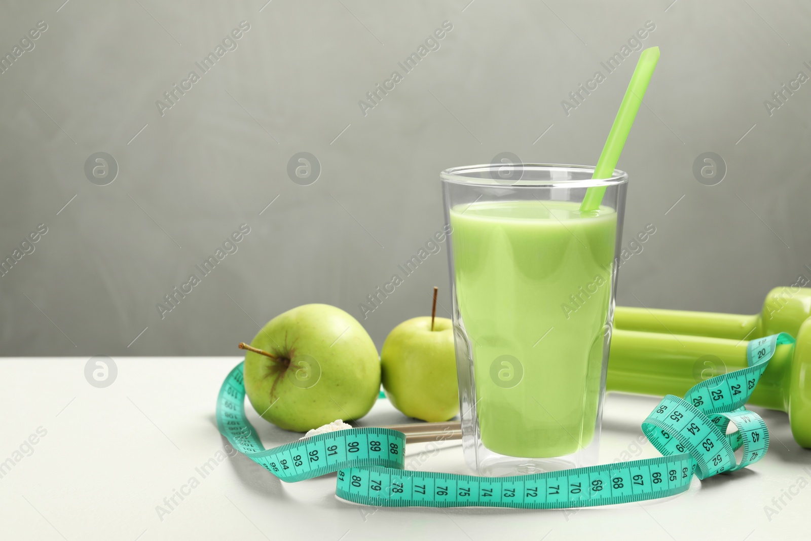 Photo of Tasty shake with apples, dumbbells and measuring tape on white table against gray background, space for text. Weight loss