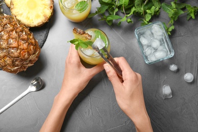 Woman putting ice in glass with cocktail  at grey table, top view