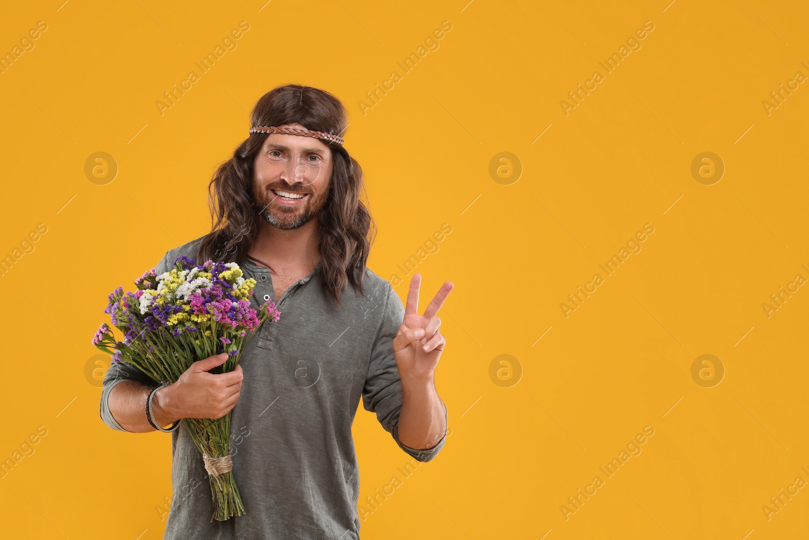 Photo of Hippie man with bouquet of colorful flowers showing V-sign on orange background, space for text