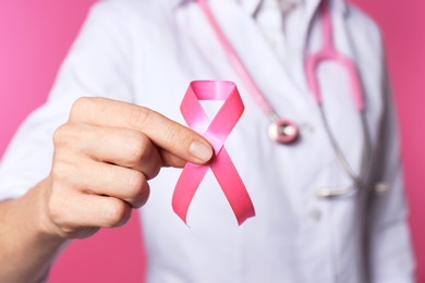 Female doctor with pink ribbon and stethoscope on color background, closeup. Breast cancer concept