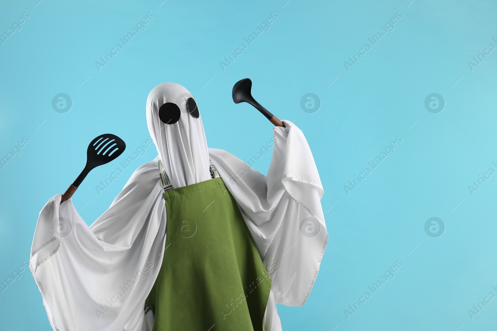 Photo of Woman in ghost costume and apron with kitchen set on light blue background, space for text