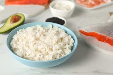 Photo of Cooked rice in bowl and other ingredients for sushi on white marble table, closeup