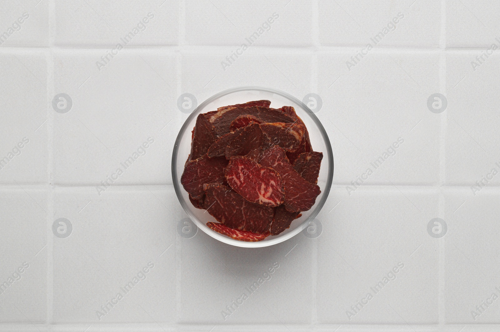 Photo of Bowl with pieces of delicious beef jerky on white tiled table, top view