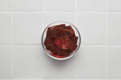 Photo of Bowl with pieces of delicious beef jerky on white tiled table, top view