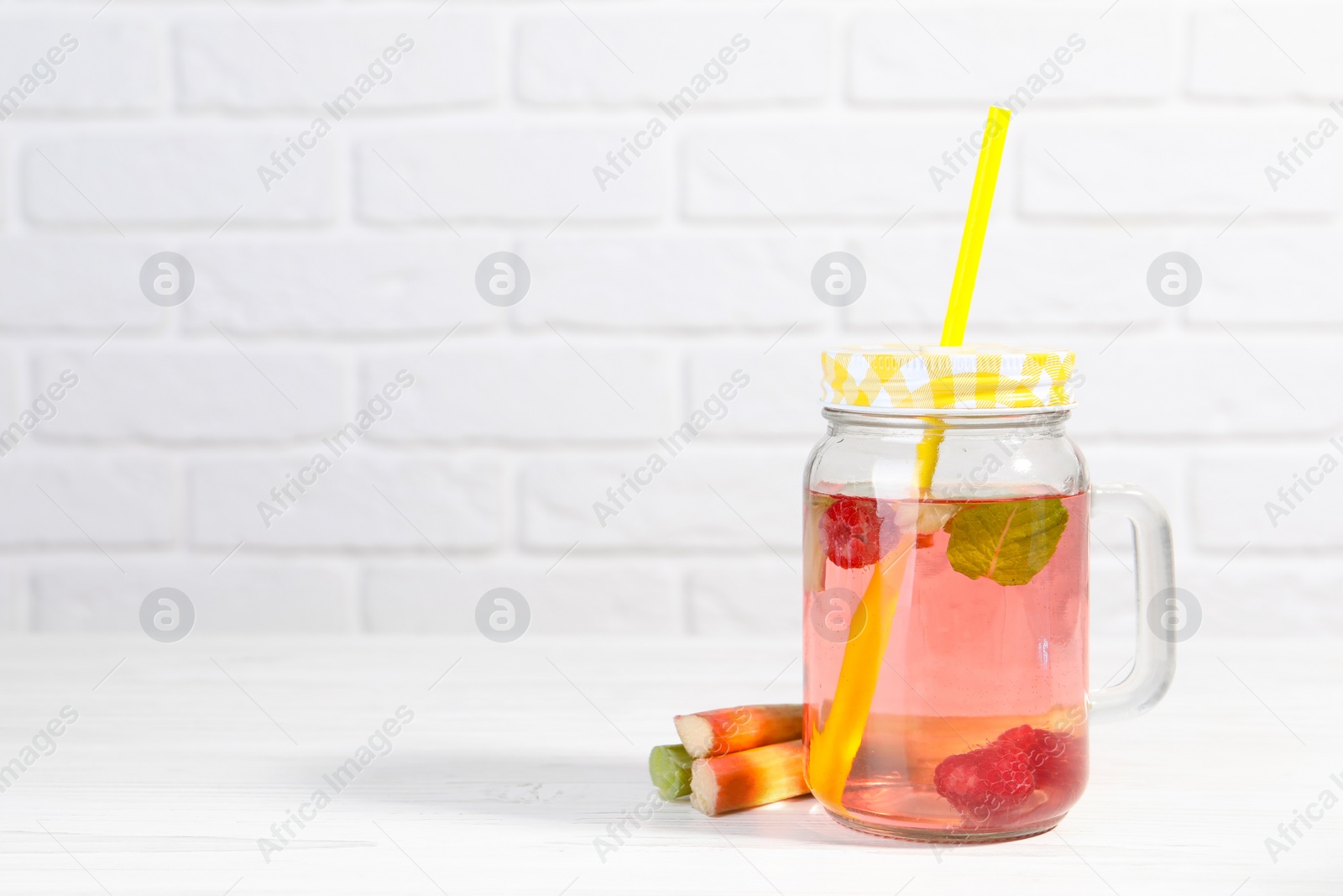 Photo of Mason jar of tasty rhubarb cocktail with raspberry and stalks on white wooden table. Space for text