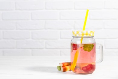 Photo of Mason jar of tasty rhubarb cocktail with raspberry and stalks on white wooden table. Space for text
