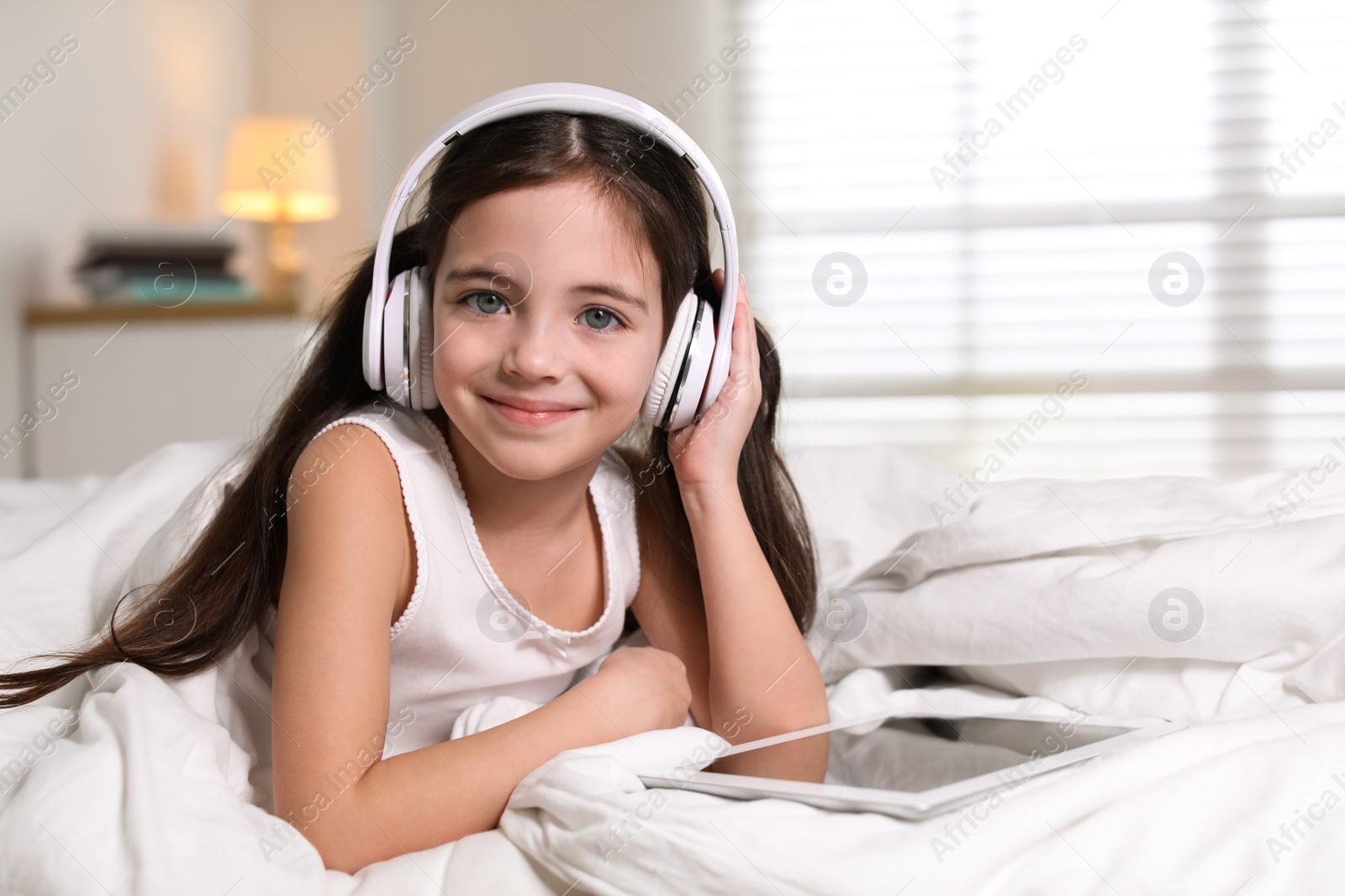 Photo of Cute little girl with headphones and tablet listening to audiobook in bed at home