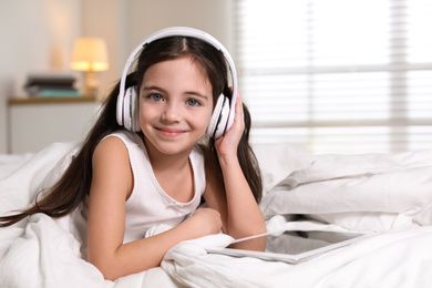 Cute little girl with headphones and tablet listening to audiobook in bed at home