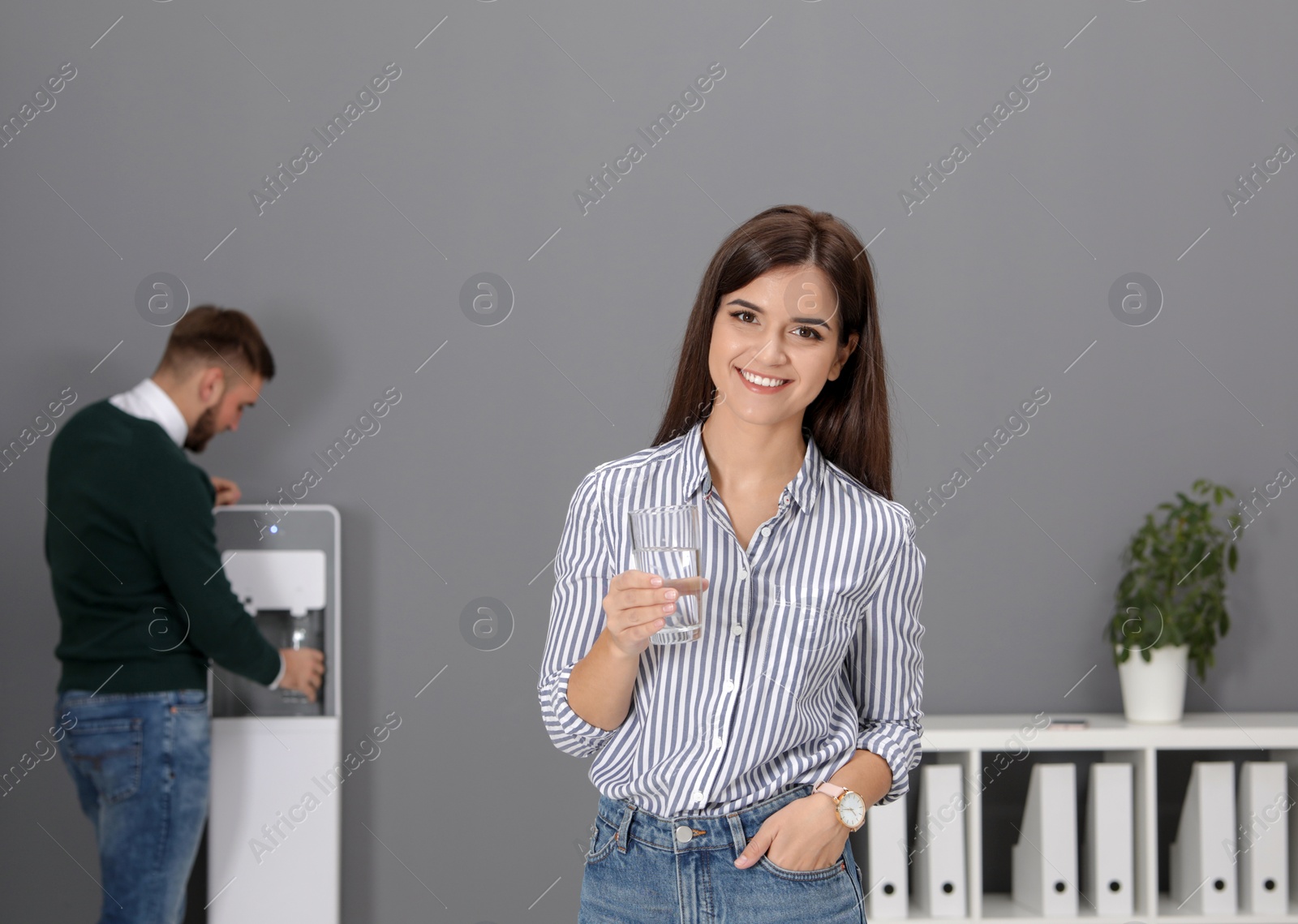 Photo of Young woman with glass of water near cooler in office