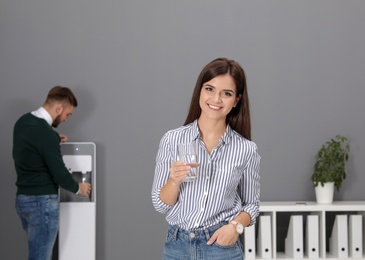 Young woman with glass of water near cooler in office